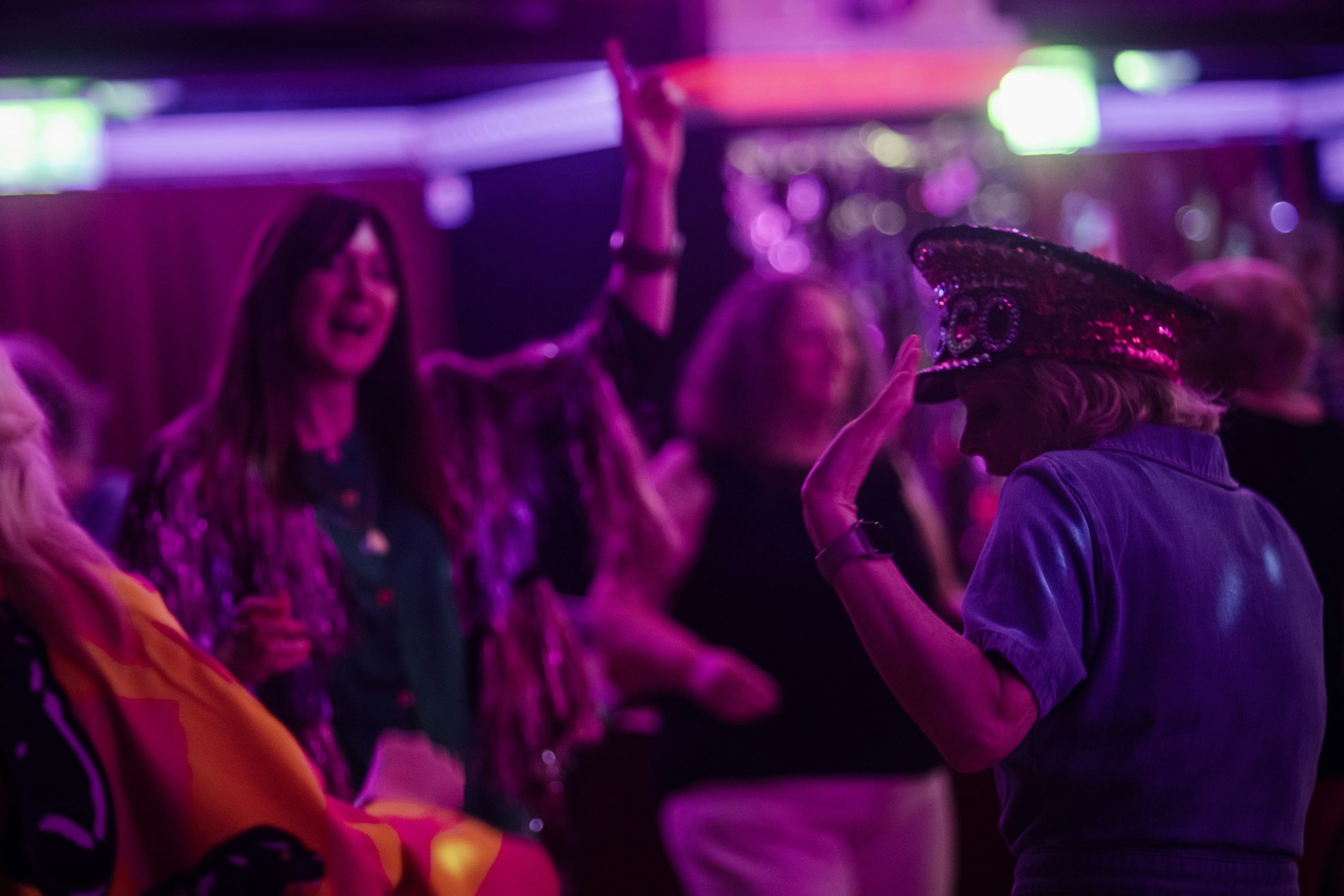 People dance under pink and purple lights at a Grief Disco, one woman wearing a sequinned disco hat